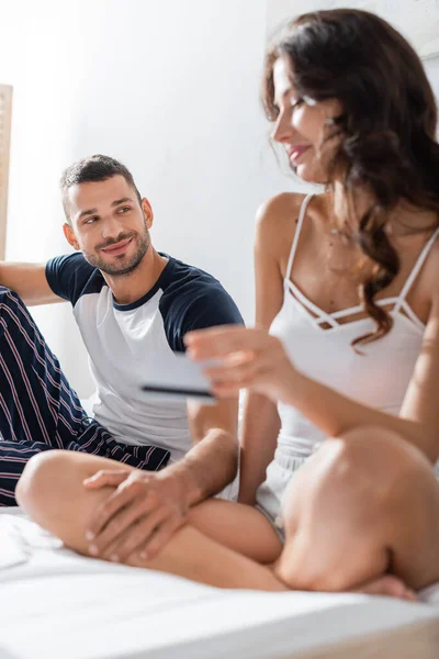 Smiling man in pajama looking at blurred girlfriend with credit card on bed — Stock Photo