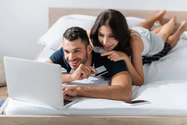 Весела жінка вказує на ноутбук біля хлопця з кредитною карткою на ліжку — стокове фото