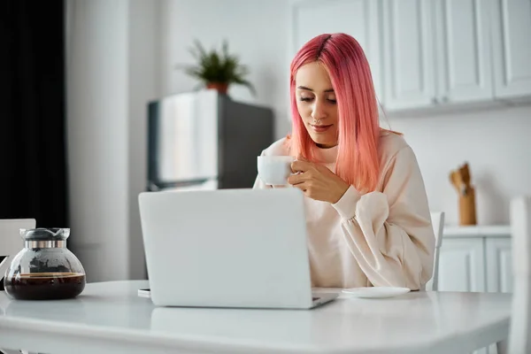 Boa aparência mulher alegre com cabelo rosa vibrante sentado no laptop e beber café na cozinha — Fotografia de Stock