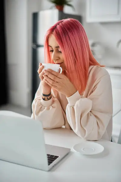 Attractive young woman with pink hair in casual attire working at laptop at home and drinking coffee — Stock Photo