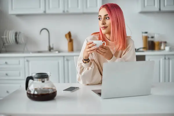 Jolie femme joyeuse avec des cheveux roses en tenue décontractée travaillant à l'ordinateur portable à la maison et buvant du café — Photo de stock