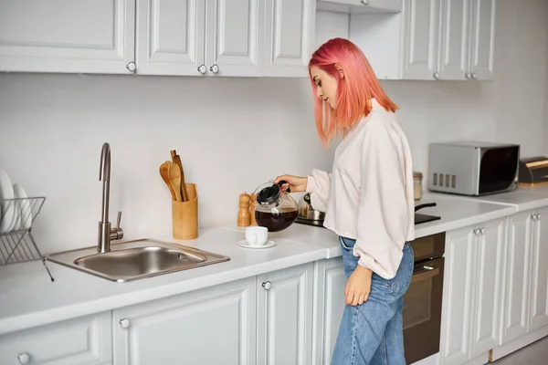 Красивая молодая женщина в повседневной одежде с розовыми волосами наливая себе кофе на кухне — стоковое фото