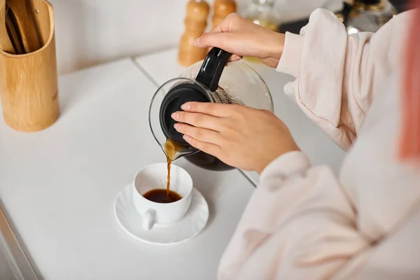 Cropped view of young woman in everyday clothes pouring hot coffee into cup at home — Stock Photo