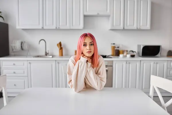 Bella giovane donna con i capelli rosa seduta in cucina a tavola e guardando dritto la fotocamera — Foto stock
