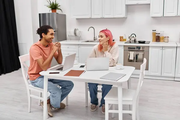 Alegre hermosa pareja interracial en ropa de casa trabajando en sus computadoras portátiles y sonriendo el uno al otro - foto de stock