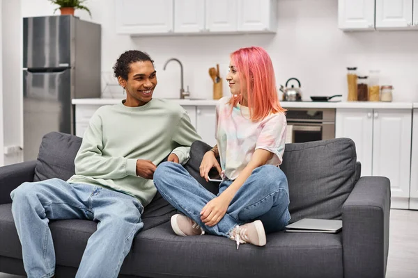 Attrayant couple interracial joyeux en vêtements de maison assis sur le canapé et souriant joyeusement à l'autre — Photo de stock