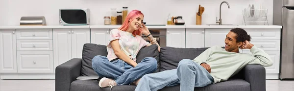 Beautiful jolly interracial couple relaxing on sofa in living room and smiling at each other, banner — Stock Photo