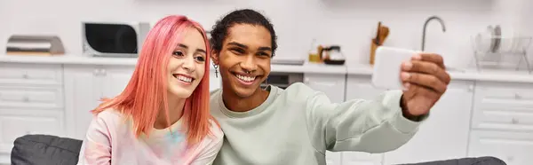 Alegre pareja multicultural atractiva tomando selfies mientras en la sala de estar en casa, pancarta - foto de stock