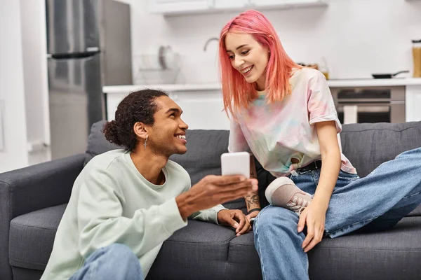 Schönes freudiges multirassisches Paar in Homewear, das im Wohnzimmer sitzt und glücklich auf das Telefon schaut — Stockfoto