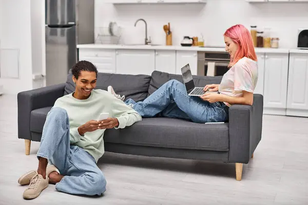 Homem americano africano alegre olhando para o telefone ao lado de sua namorada alegre trabalhando no laptop — Fotografia de Stock