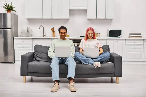 Joyeux attrayant couple interracial en vêtements de maison assis sur le canapé et travaillant sur des ordinateurs portables à distance — Photo de stock