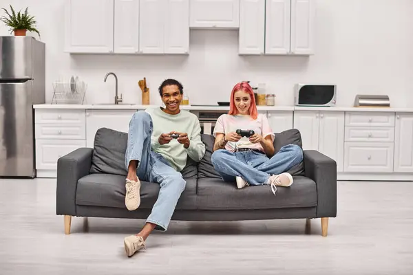 Alegre interracial casal no homewear sentado no sofá e jogar jogos usando gamepads em casa — Fotografia de Stock