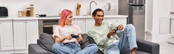 Alegre casal multirracial em trajes confortáveis sentado e usando gamepads para jogar jogos na sala de estar — Fotografia de Stock