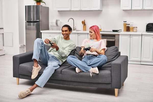 Jolly multicultural couple in cozy homewear sitting and playing video games with joysticks at home — Stock Photo