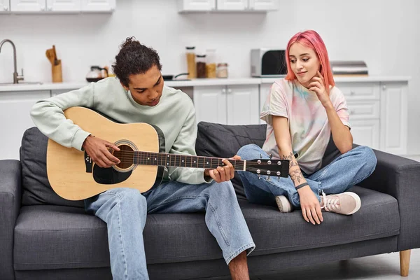 Alegre rosa cabelo bonita mulher olhando para seu namorado bonito tocando guitarra na sala de estar — Fotografia de Stock