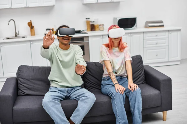 Cheerful appealing multicultural couple sitting on sofa in living room wearing VR headsets — Stock Photo