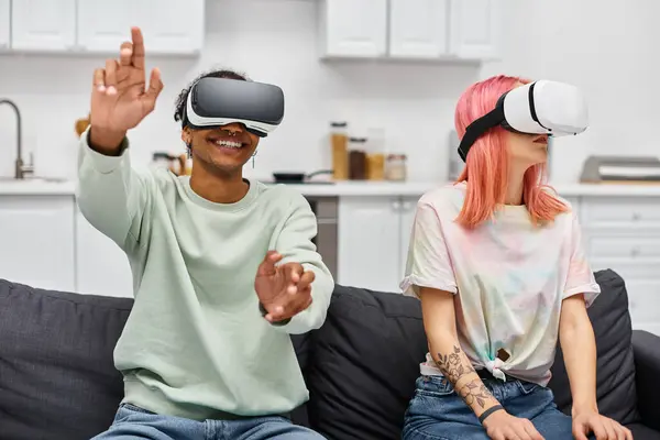 Joyous beautiful multiracial couple in homewear sitting on sofa with virtual reality headsets — Stock Photo