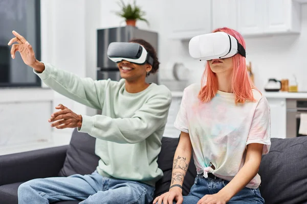 Joyful attractive diverse couple in casual attire sitting on sofa in living room with VR headsets — Stock Photo