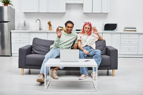 Joyful beautiful multicultural couple sitting on sofa and waving at laptop camera during video call — Stock Photo
