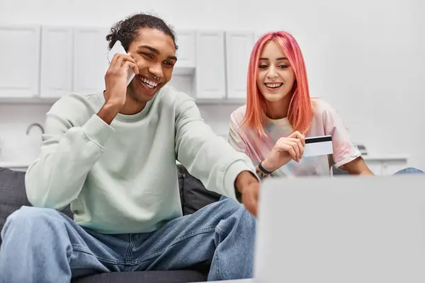 Appealing jolly diverse couple looking at laptop while holding credit card and talking by phone — Stock Photo