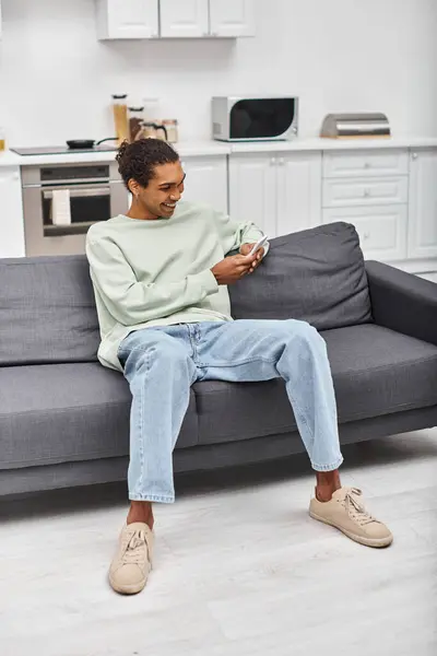 Bel homme heureux afro-américain dans des vêtements confortables assis sur le canapé et regardant le téléphone — Photo de stock