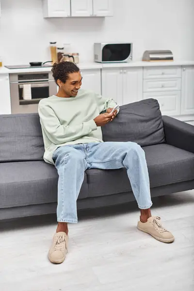 Handsome joyous african american man in comfy homewear sitting on sofa and looking at phone — Stock Photo