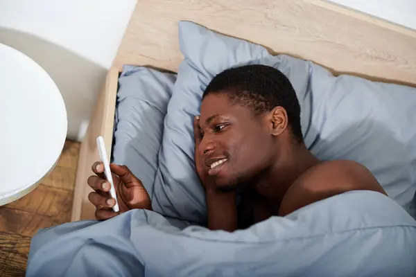 Young African American man laying in bed, holding smartphone in his hand — Stock Photo