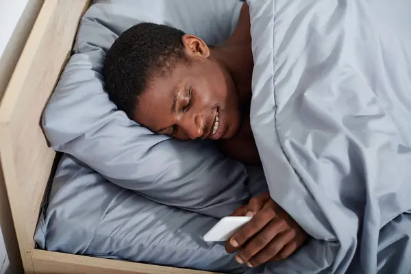 A young African American man peacefully laying in bed, holding smartphone while waking up in the morning. — Stock Photo