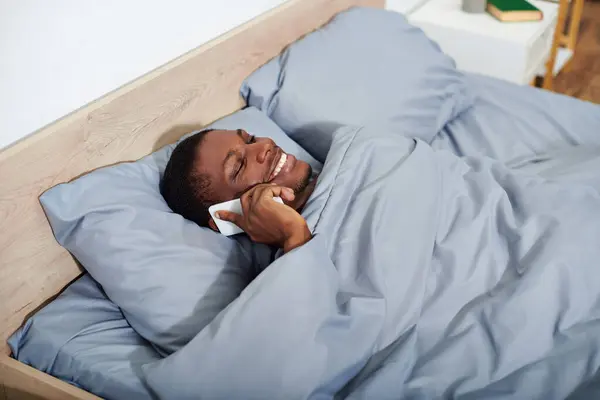 A young African American man laying in bed, holding a smartphone in his hand as he wakes up in the morning. — Stock Photo