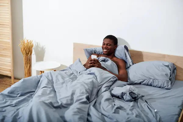 A young African American man lays in bed, fixated on his cell phone screen — Stock Photo