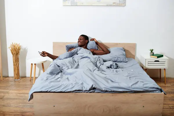 Young African American man laying in bed, holding a cell phone, waking up in the morning. — Stock Photo
