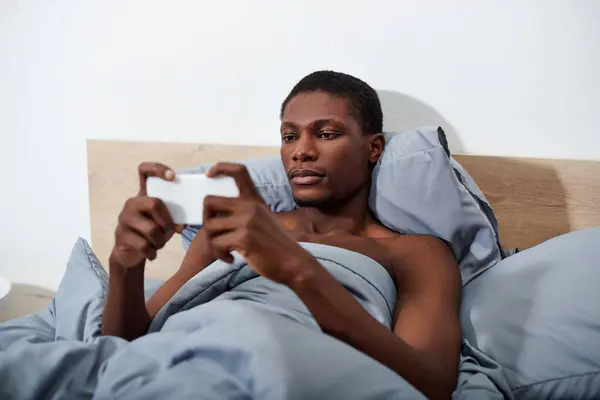 A young African American man relaxes in bed, holding a cell phone in his hands. — Stock Photo