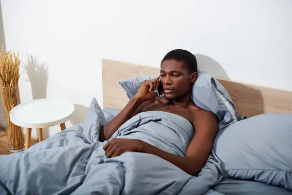 An African American man lays in bed, chatting on his cell phone in the morning, starting his day off with a phone call. — Stock Photo