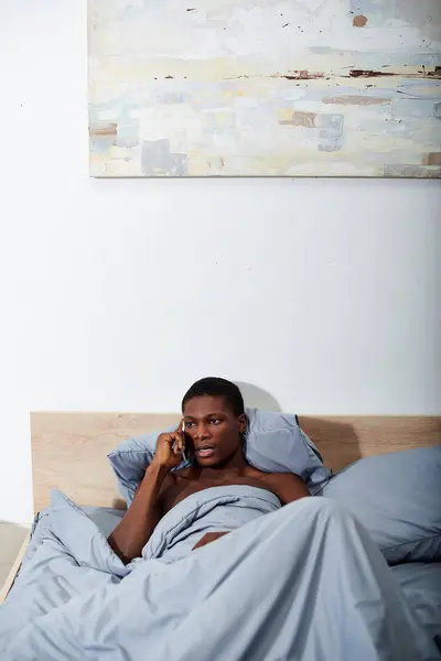 A young African American man lays in bed, talking on a cell phone in the morning light. — Stock Photo