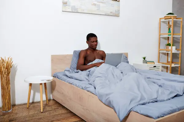 African American man sitting on bed, focused on laptop screen early in the morning. — Stock Photo