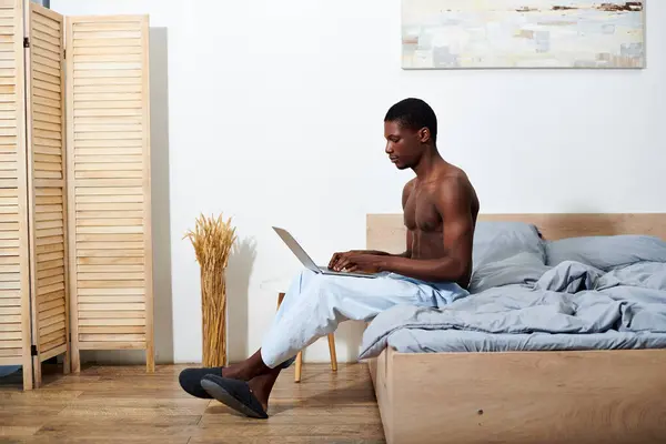 A young African American man sits on a bed, engrossed in using a laptop computer. — Stock Photo