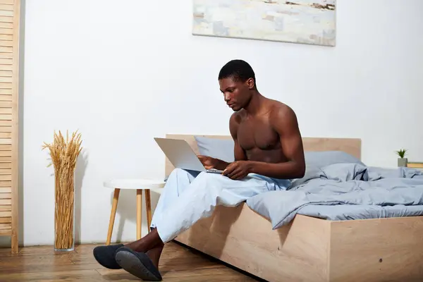 Ein junger afroamerikanischer Mann sitzt auf einem Bett und konzentriert sich auf die Benutzung eines Laptops. — Stockfoto