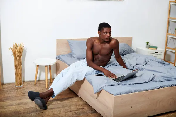 Young African American man sits on bed, focusing on his laptop screen in the morning. — Stock Photo