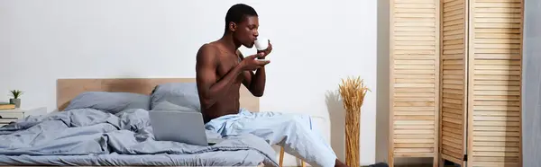 A young African American man, seated on a bed, operates a laptop with intense focus. — Stock Photo