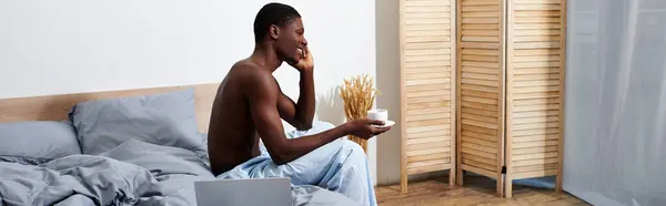 A young African American man sits on a bed, holding a cup of coffee, enjoying a peaceful morning moment. — Stock Photo