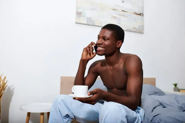 Young African American man sits on bed in morning, engrossed in conversation on cell phone. — Stock Photo