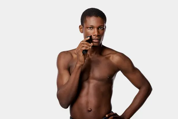 An African American man with no shirt is shaving with electric razor in a studio with a white background. — Stock Photo