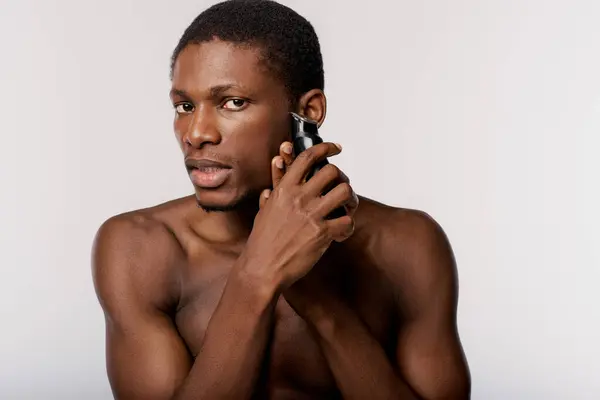 Shirtless African American man shaving with electric razor in a modern studio setting against a white background. — Stock Photo