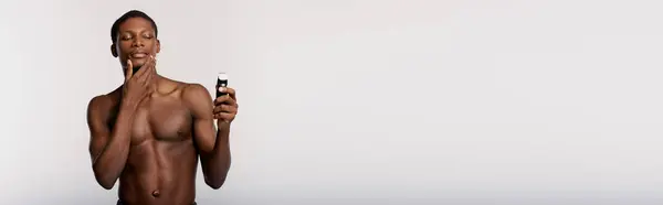Shirtless African American man shaving with electric razor against a white background, showcasing his daily beauty routine. — Stock Photo