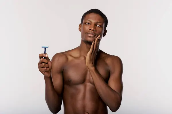 A shirtless African American man holds a razor in his right hand in a studio with a white background. — Stock Photo