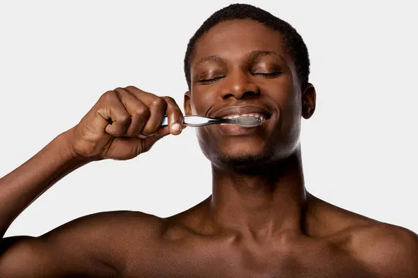 An African American man, shirtless, brushing his teeth with a toothbrush against a white background. — Stock Photo