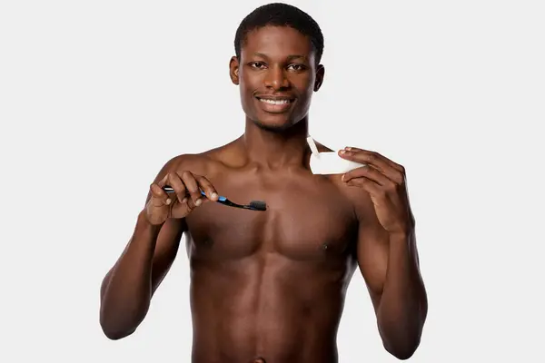 A shirtless African American man holds a toothbrush and toothpaste in his right hand against a white background. — Stock Photo