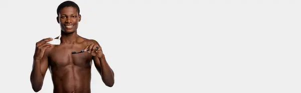 Безрубашечный афроамериканец держит зубную щетку и зубную пасту в правой руке. — стоковое фото
