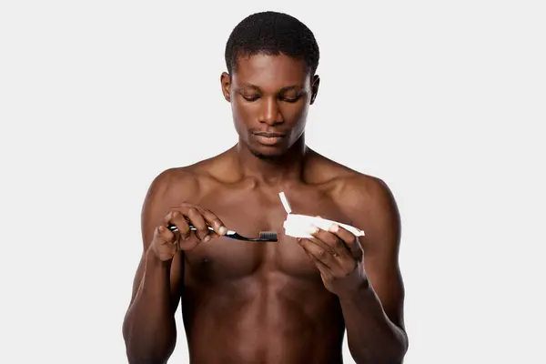 Uomo afroamericano senza camicia tiene spazzolino da denti in una mano e dentifricio nell'altra, in un ambiente studio con sfondo bianco. — Foto stock