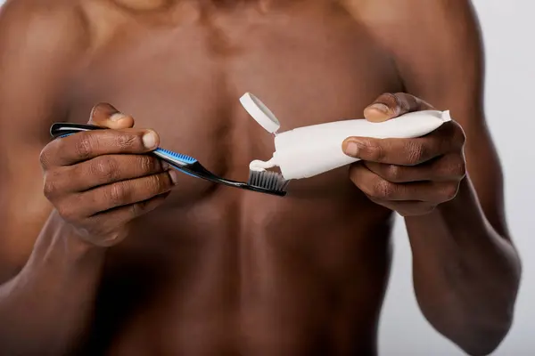 Un hombre afroamericano sosteniendo un cepillo de dientes y pasta de dientes en un estudio con un fondo blanco. - foto de stock